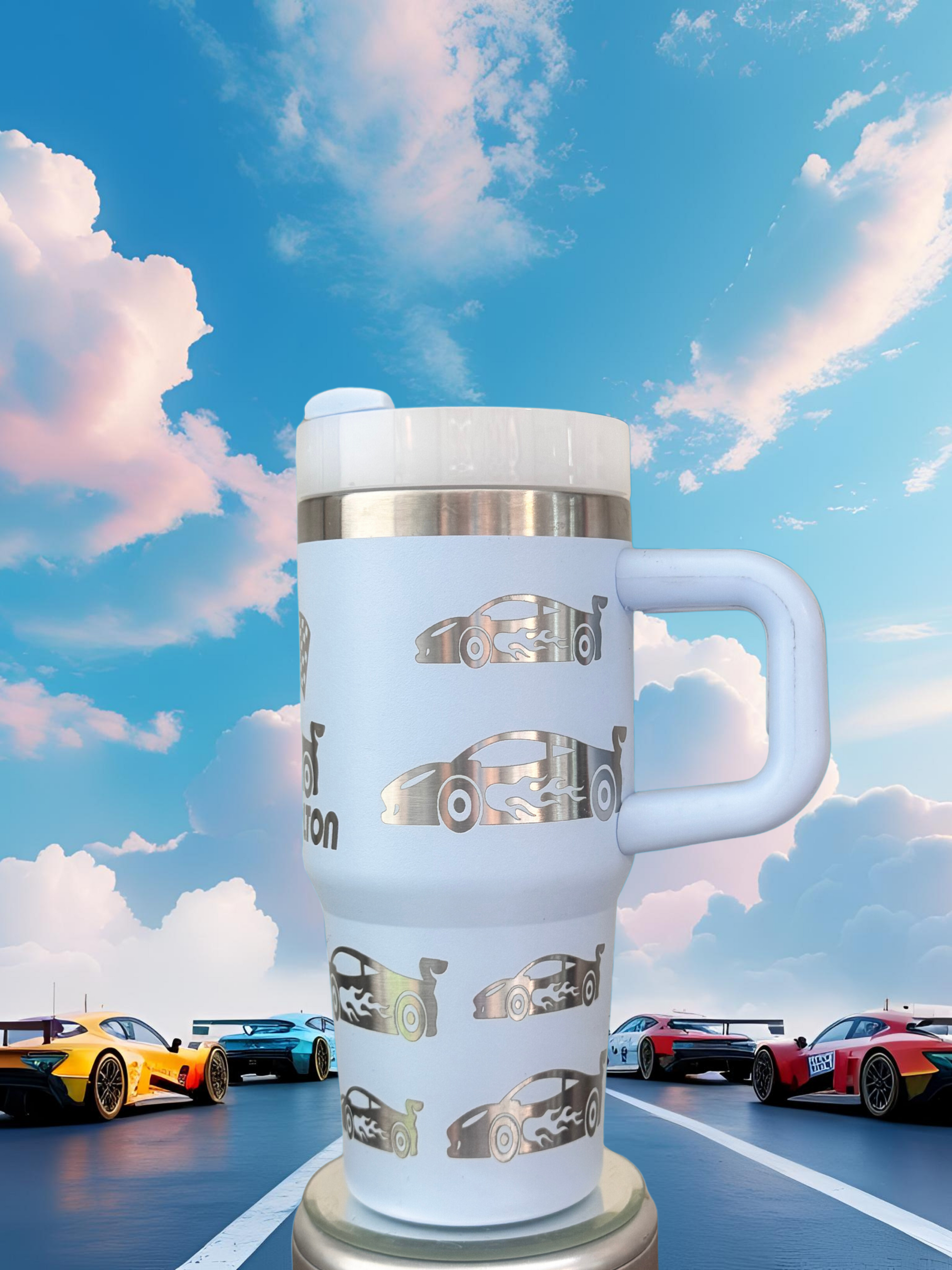 A white thermos mug with a handle and lid, featuring a design of race cars, is placed in the foreground against a backdrop of colorful race cars on a racetrack under a blue sky with clouds.