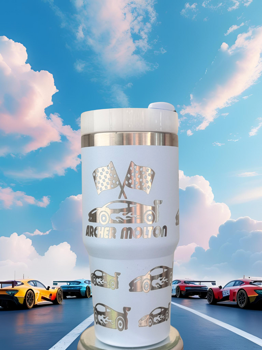 A white tumbler with a silver lid, featuring a design of racing cars and checkered flags, is placed in the foreground against a backdrop of a cloudy blue sky and racing cars.